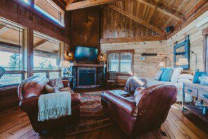 Thayne Wyoming getaway showing a rustic, cozy cabin interior featuring a warm fireplace, leather armchairs, wooden vaulted ceilings, and large windows looking out at scenic views.