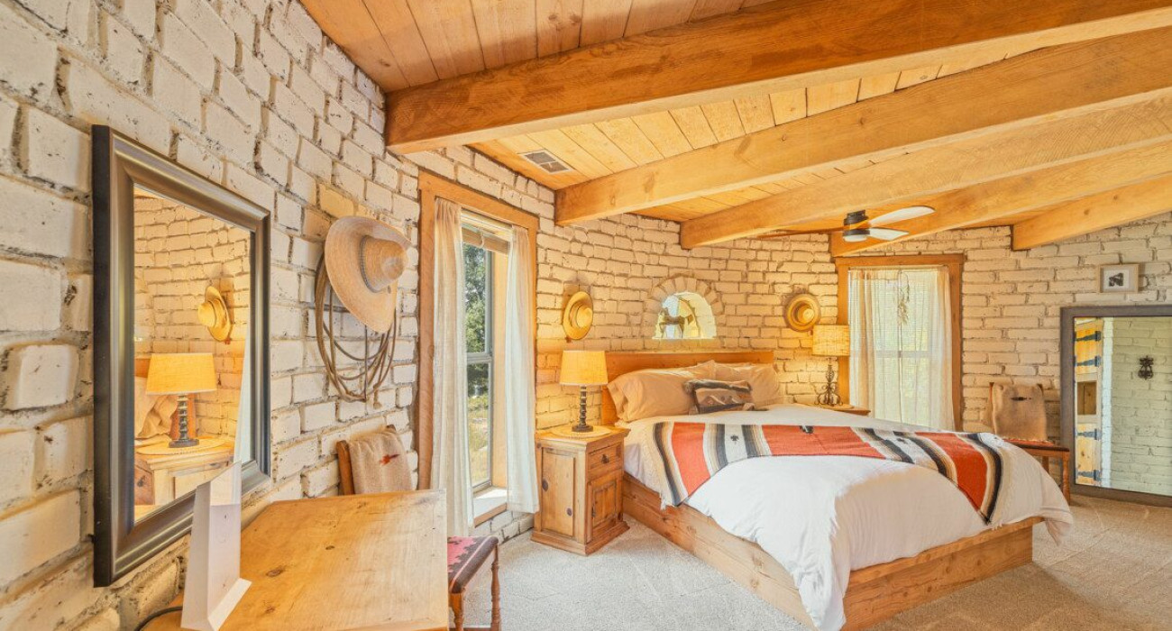 A cozy bedroom featuring a bed and a mirror. The interior design showcases hardwood flooring and wooden beams on the ceiling, creating a warm atmosphere. This space is part of the Coarsegold home rental, Casa de Oro.