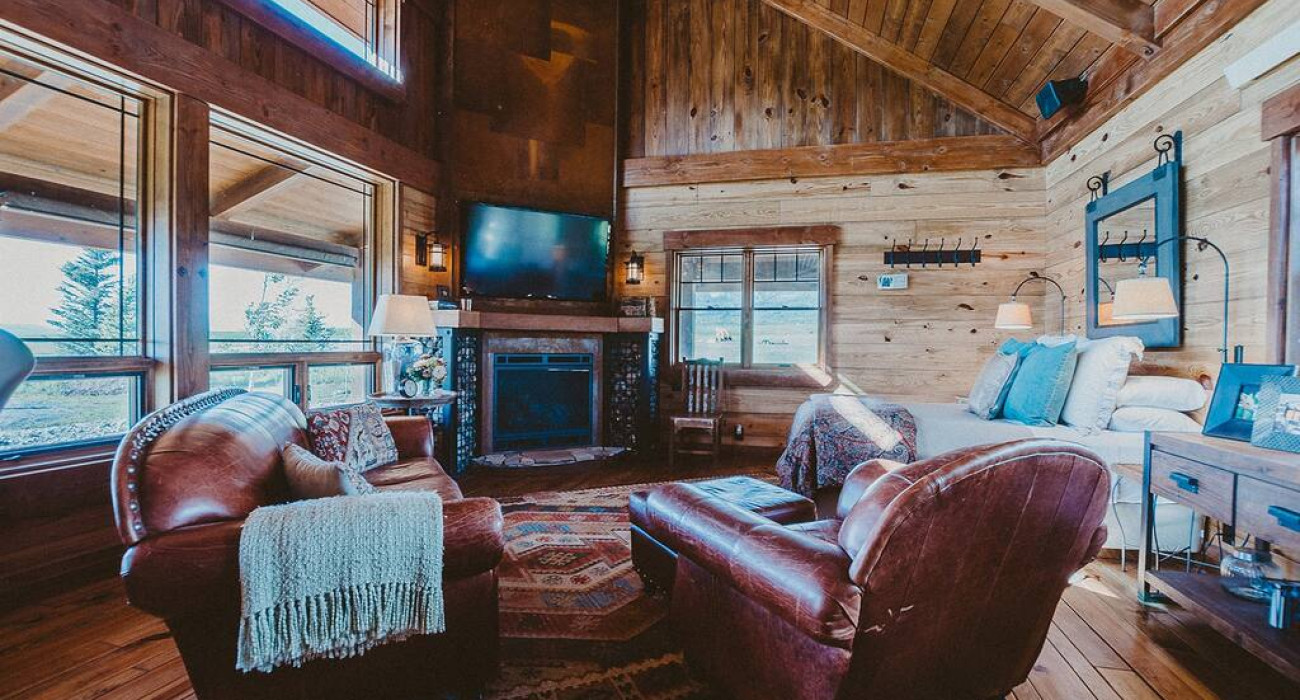 Thayne Wyoming getaway showing a rustic, cozy cabin interior featuring a warm fireplace, leather armchairs, wooden vaulted ceilings, and large windows looking out at scenic views.